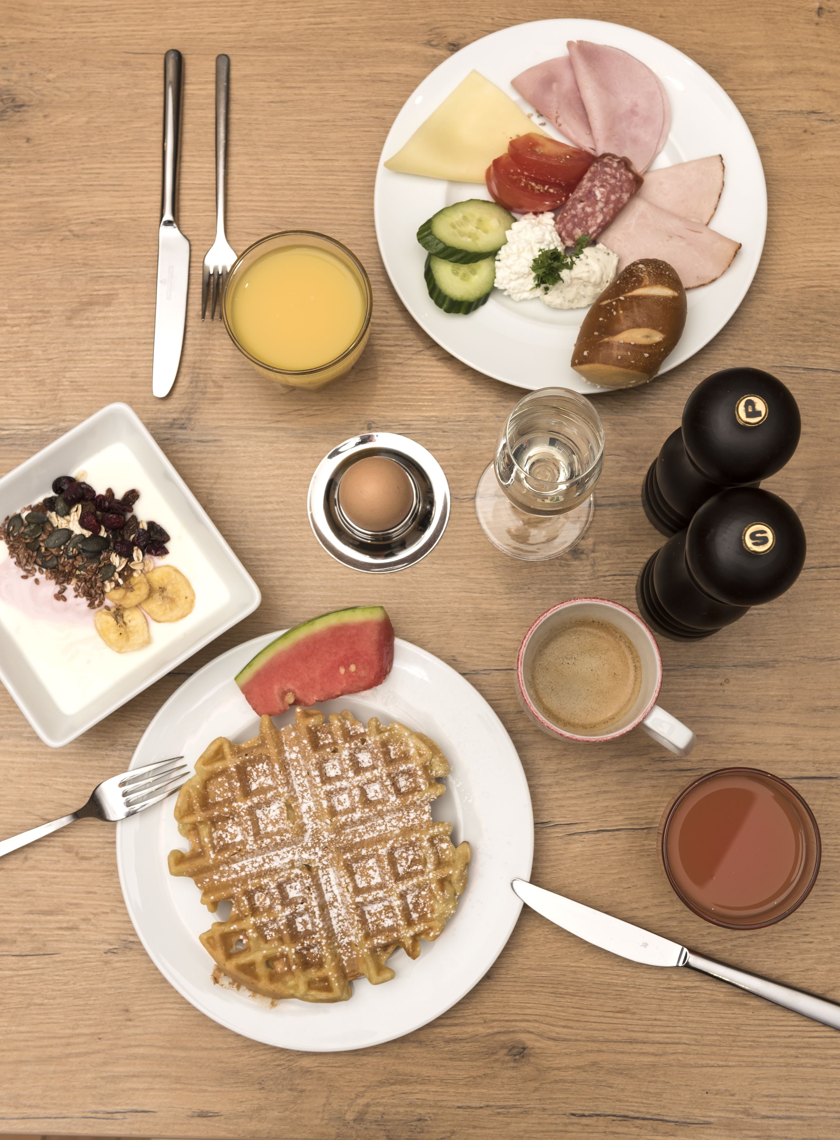A breakfast setting with waffles, ham, cheese, tomatoes, cucumber, and a slice of watermelon on a wooden table.