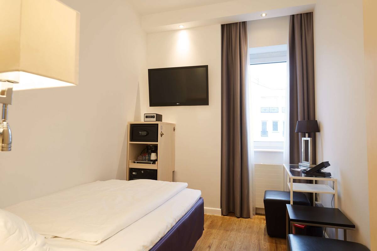 A hotel room with a bed, a desk, a flat-screen TV, a lamp, a chair, and a window with curtains.
