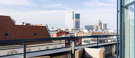 Una terraza en la azotea con muebles de mimbre, una mesa de vidrio y vistas al horizonte urbano que incluye edificios modernos e históricos.