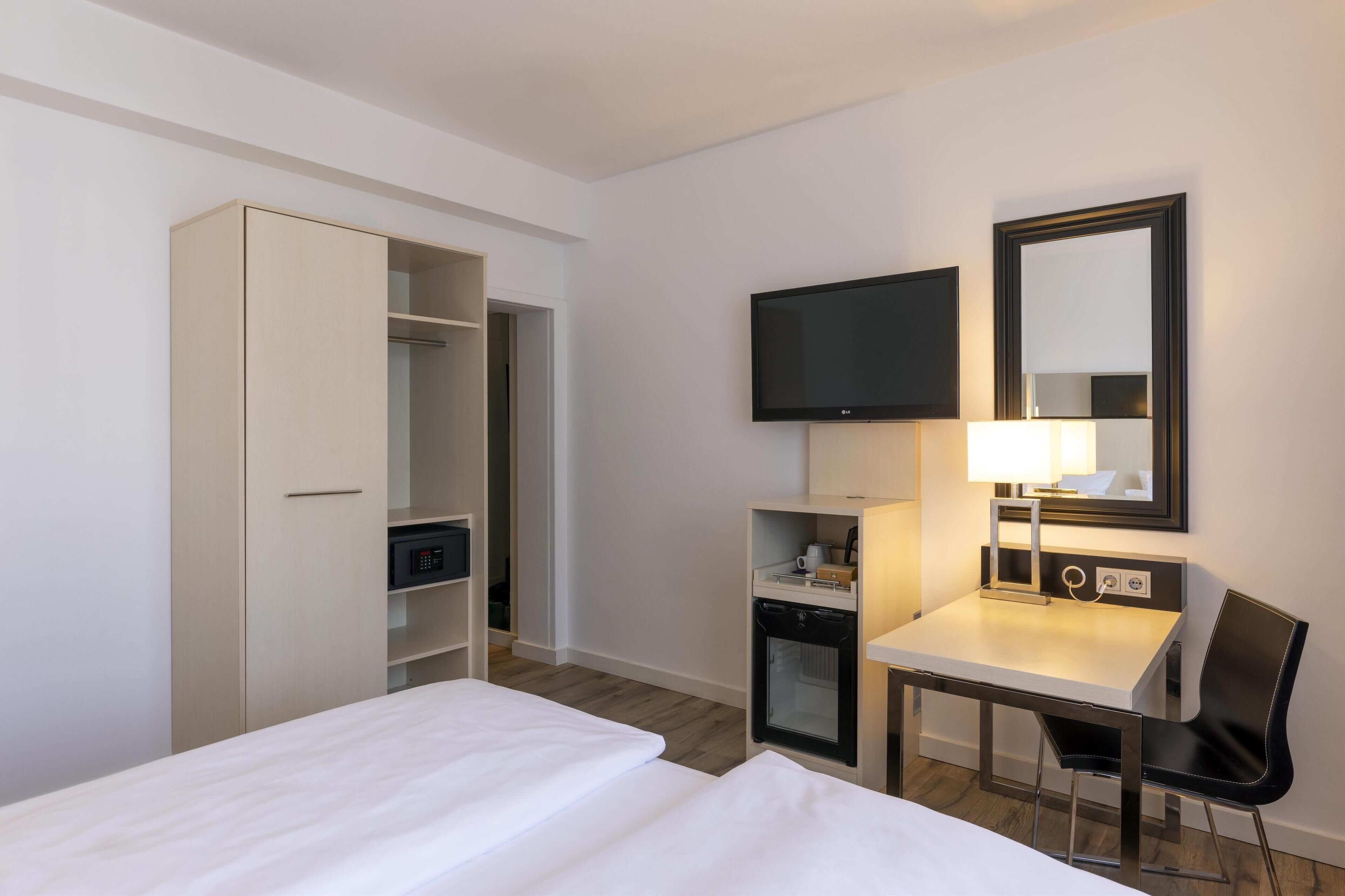 Habitación de hotel con cama, escritorio con silla, televisor, espejo y armario.