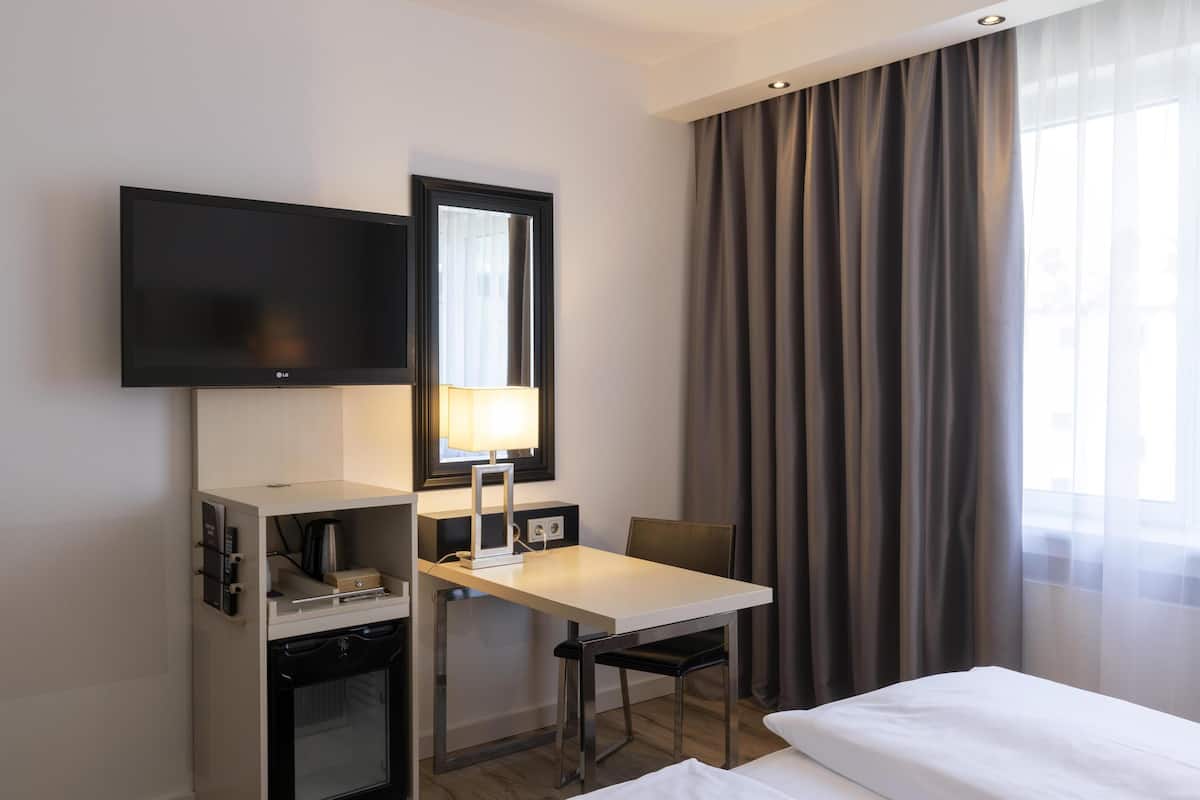A hotel room with a flat-screen TV, a desk with a lamp, a chair, a bed, and a window with curtains.