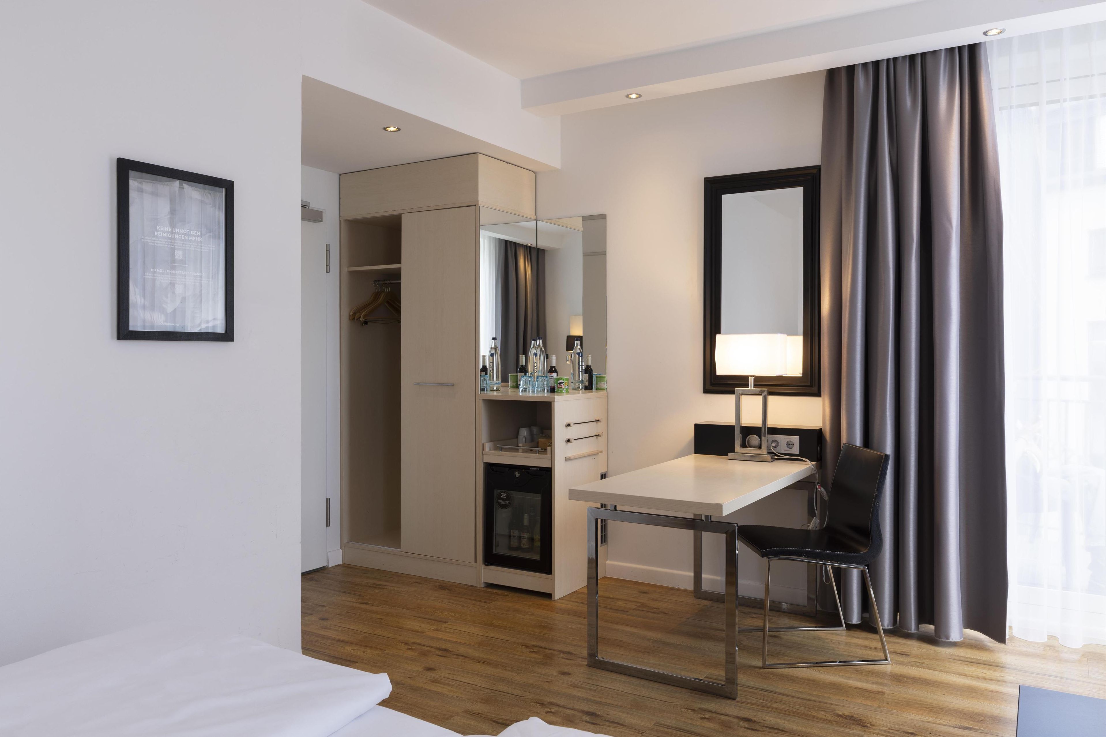 A hotel room with a bed, a desk with a chair, a wardrobe, a mirror, and a window with curtains.