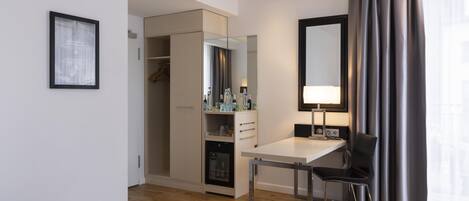 A hotel room with a bed, a desk with a chair, a wardrobe, a mirror, and a window with curtains.
