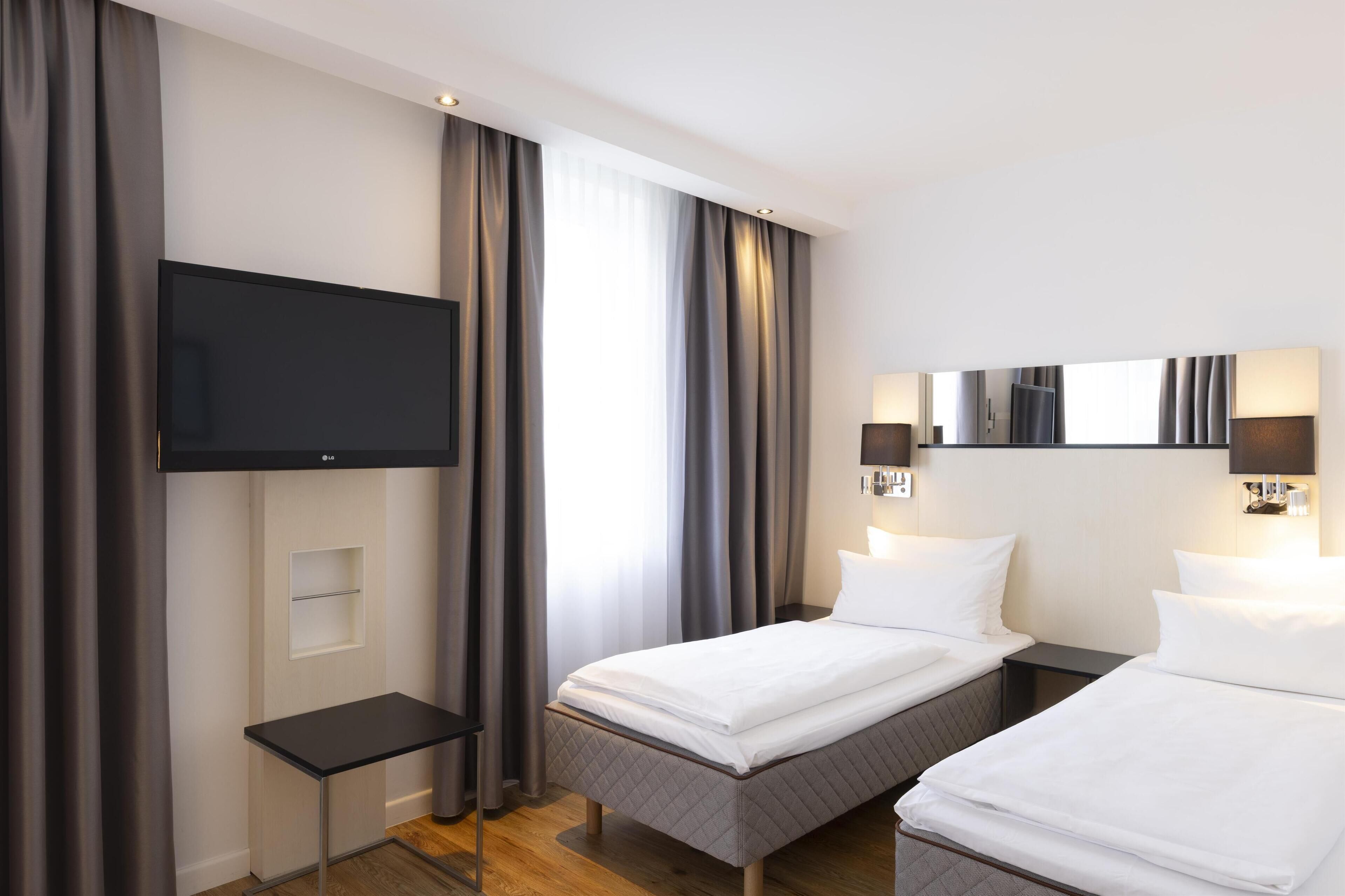 Habitación de hotel con dos camas, un televisor de pantalla plana montado en la pared, una mesita y una ventana con cortinas.