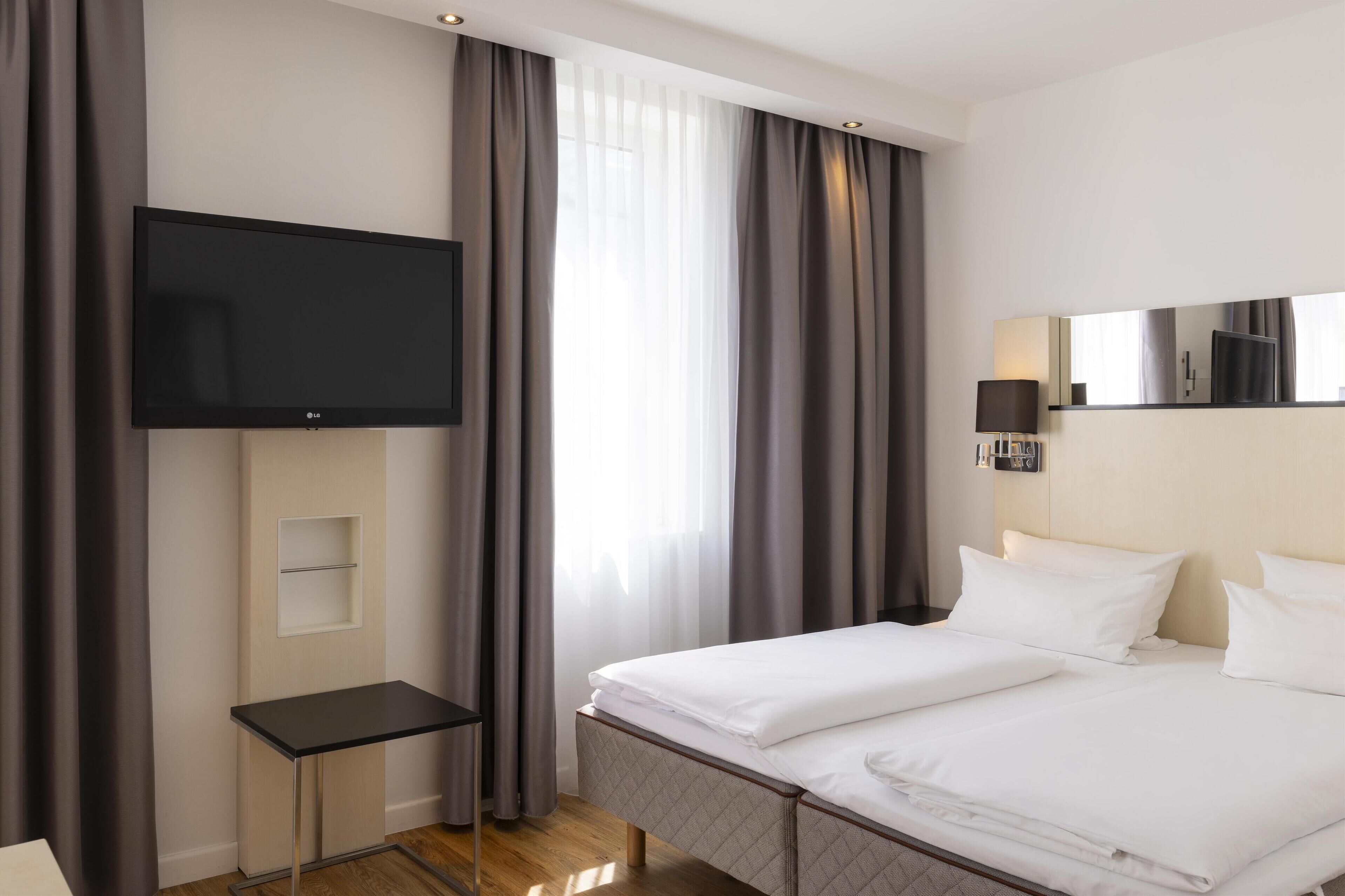 A hotel room with a bed, a TV mounted on the wall, a bedside table, and a window with curtains.