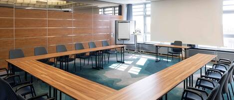 Un salón de conferencias con una gran mesa de madera, varias sillas, una pantalla de proyector y un amplio ventanal que proporciona luz natural.