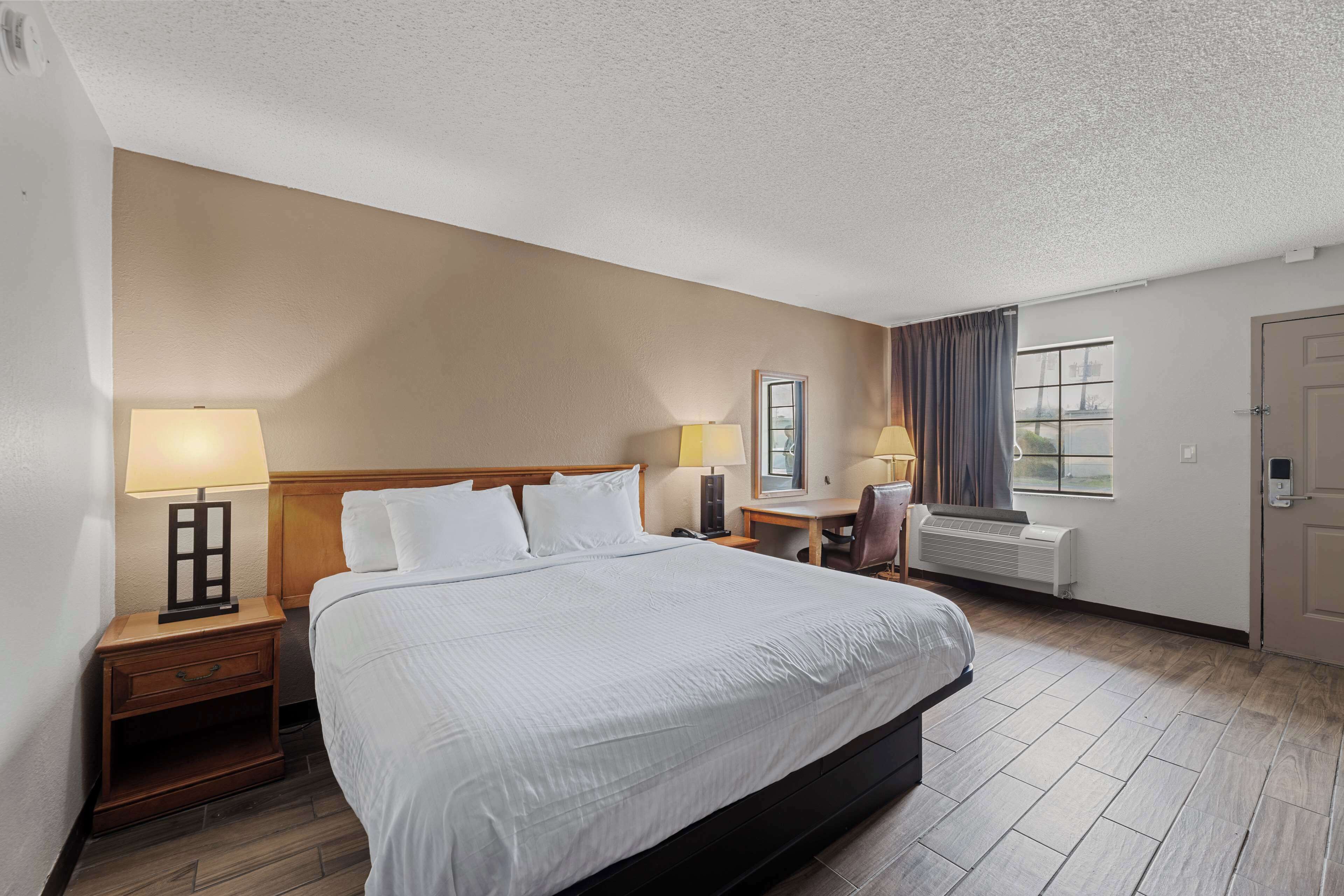 A hotel room with a large bed, bedside tables with lamps, a desk, a chair, a window with curtains, and a door.