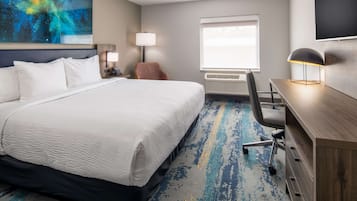 Une chambre d’hôtel avec un grand lit, un bureau, une chaise, une lampe, une télévision et une fenêtre avec des stores.