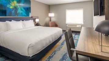 A hotel room with a large bed, a desk, a chair, a lamp, a chair, a painting, and a window.