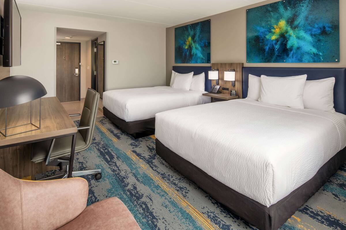 A hotel room with two beds, a desk, a chair, and a large abstract painting on the wall.