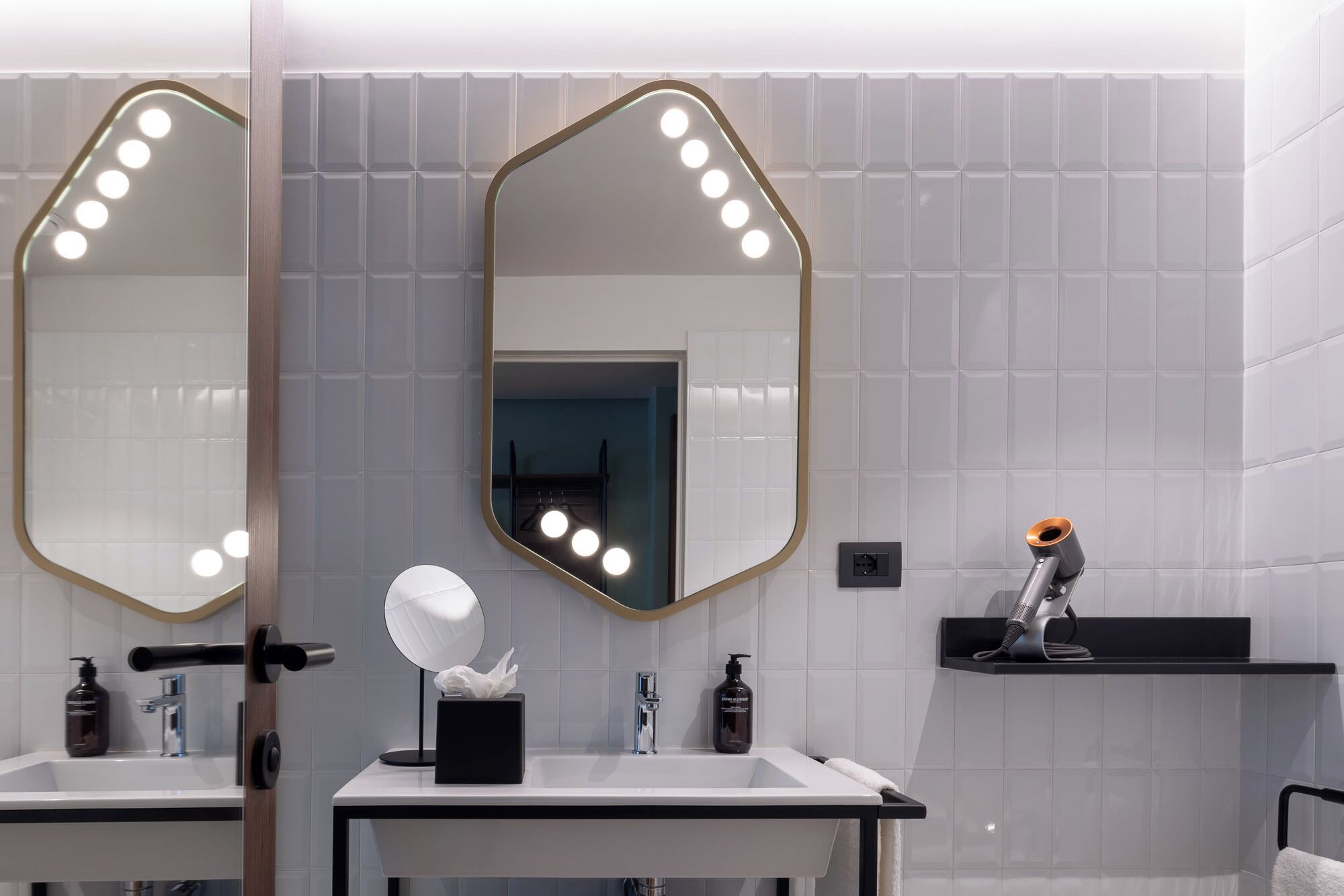 A modern bathroom with two sinks, a large mirror with lights, a towel rack, and a hairdryer on a shelf.