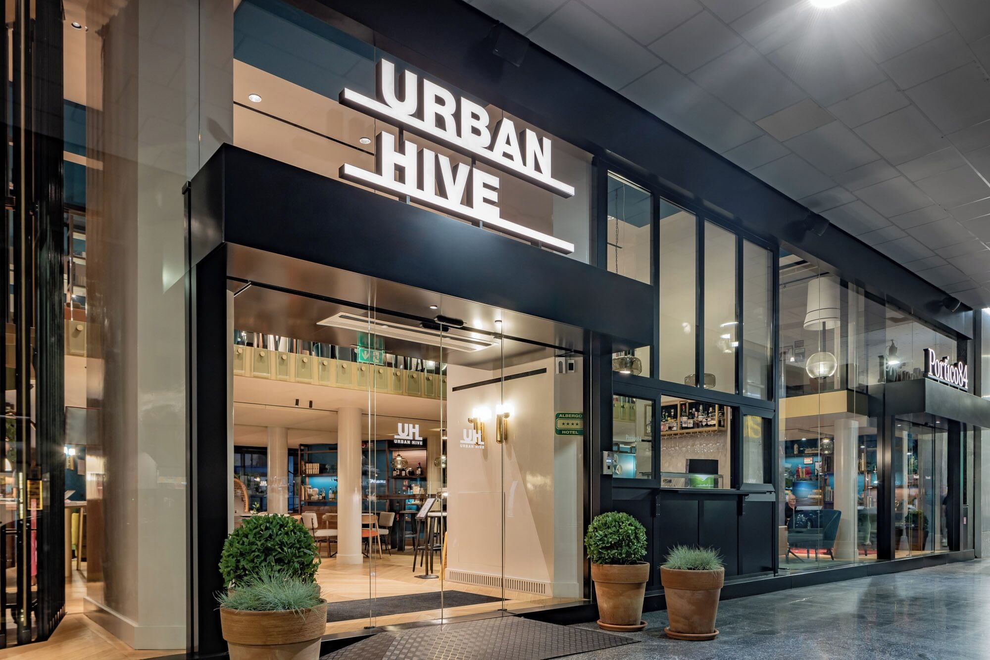 A modern hotel entrance with "URBAN HIVE" signage, glass doors, and potted plants.