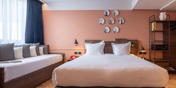 Een hotelkamer met een groot bed, een slaapbank, een wandklok, een boekenplank en een raam met gordijnen.