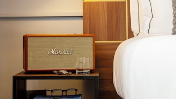A neatly arranged hotel room with a bedside table featuring a vintage Marshall speaker, a glass of water, and a stack of books. A wall-mounted lamp provides bedside lighting, and a wooden headboard with a white pillow is visible.