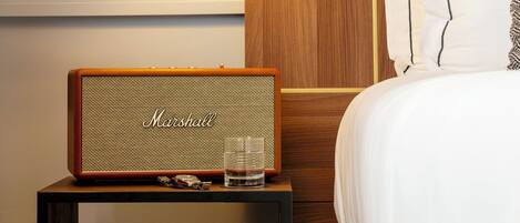 A neatly arranged hotel room with a bedside table featuring a vintage Marshall speaker, a glass of water, and a stack of books. A wall-mounted lamp provides bedside lighting, and a wooden headboard with a white pillow is visible.