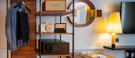 Habitación de hotel con una estantería de madera que contiene libros, un altavoz Marshall y una caja fuerte. Un espejo redondo cuelga de la pared, y una lámpara está sobre una mesita de noche.