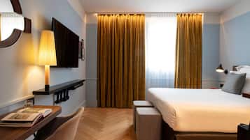 Een hotelkamer met een bed, nachtkastje, lamp en een groot raam met gordijnen.