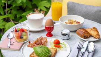 Een ontbijttafel met een croissant, toast met avocado, een schaal fruit, een glas sinaasappelsap en een kop koffie.