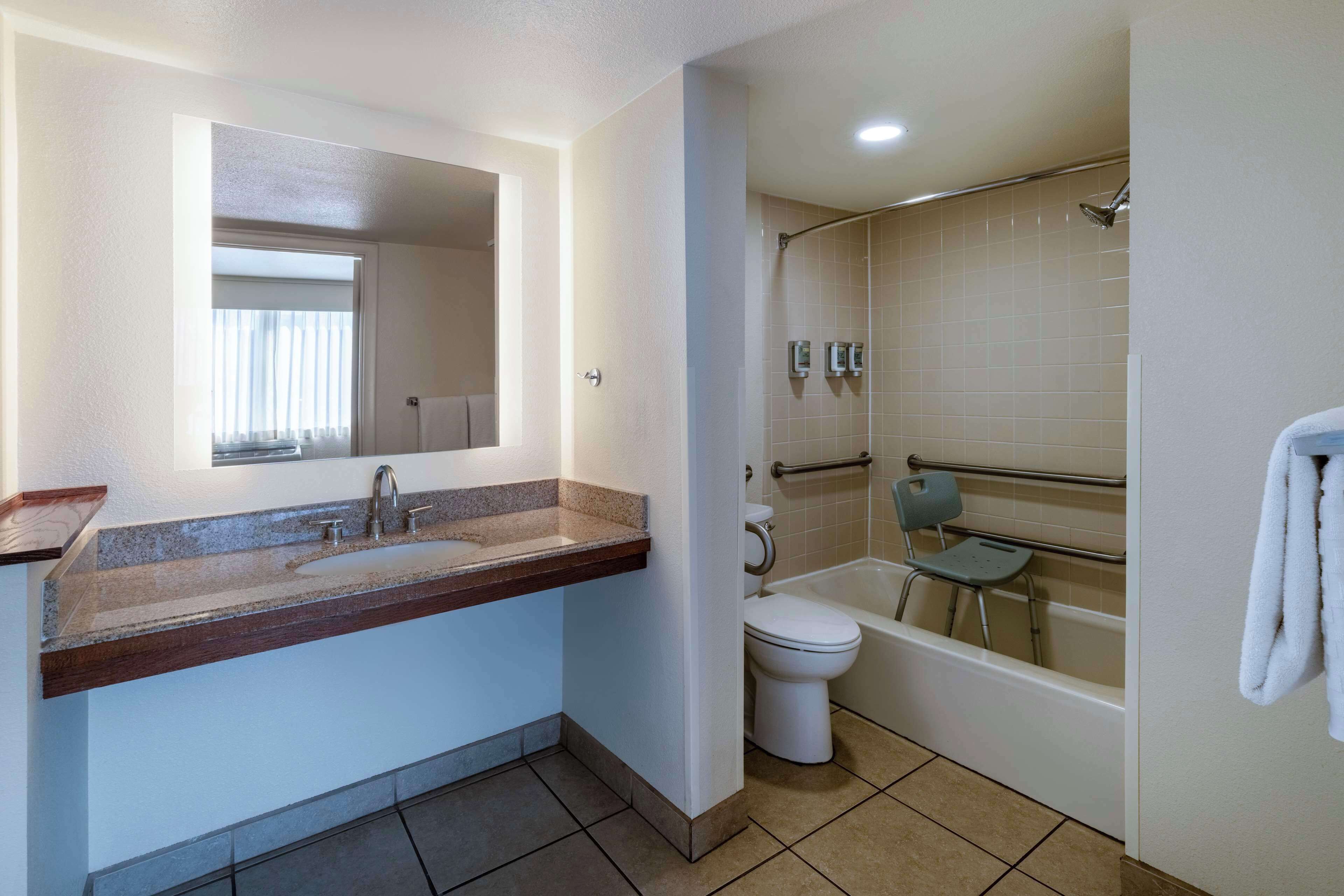 Un baño con lavamanos, inodoro y bañera, que incluye una silla de ducha y barras de apoyo.