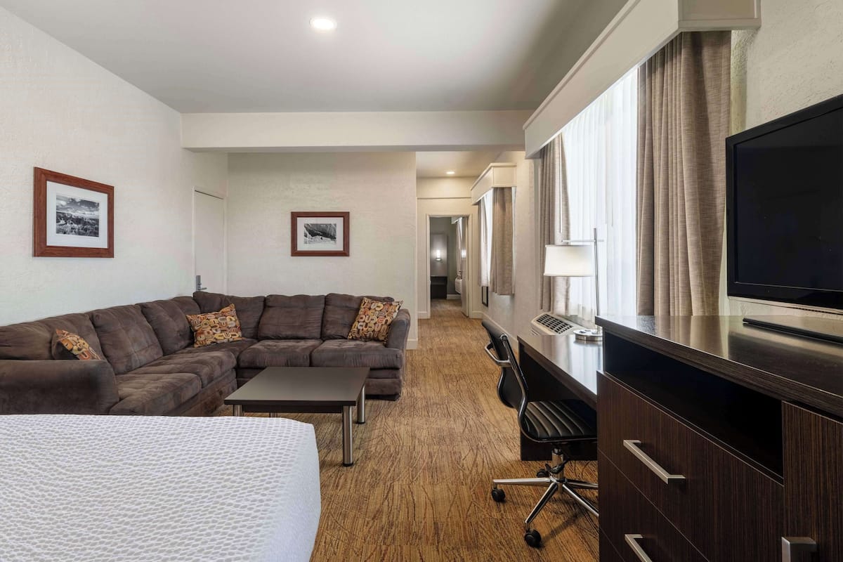 A hotel room with a large bed, a dark brown sofa, a small table, a TV, and a desk with drawers.