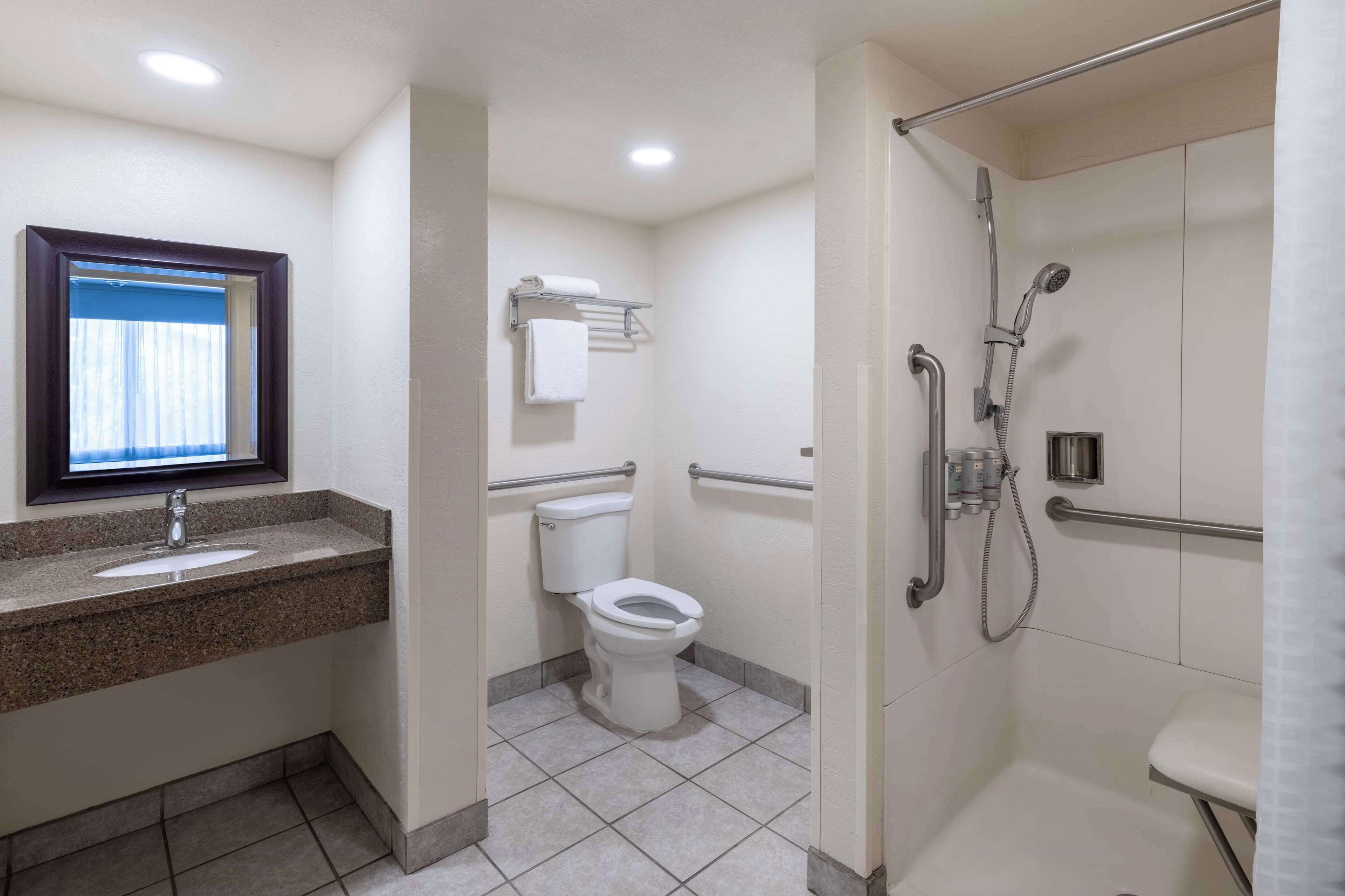Ein modernes Badezimmer mit Granitwaschtisch, Toilette, Dusche inklusive Haltegriffen und einem Fenster mit Vorhängen.