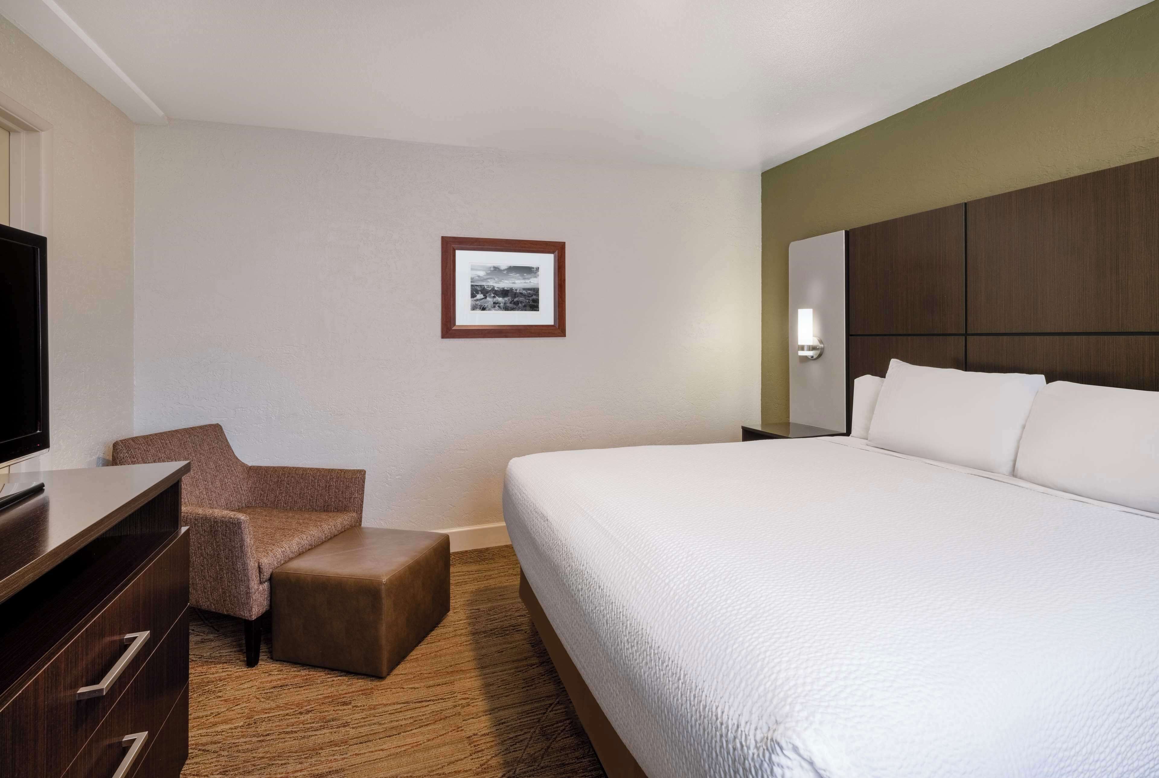 A hotel room with a large bed, a TV, a brown armchair, a small ottoman, and a framed picture on the wall.