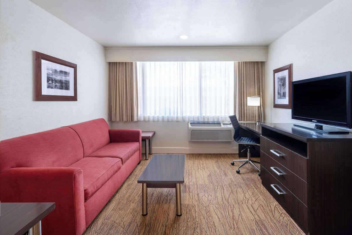 A hotel room with a red sofa, a small table, a TV on a wooden cabinet, and a large window with curtains.