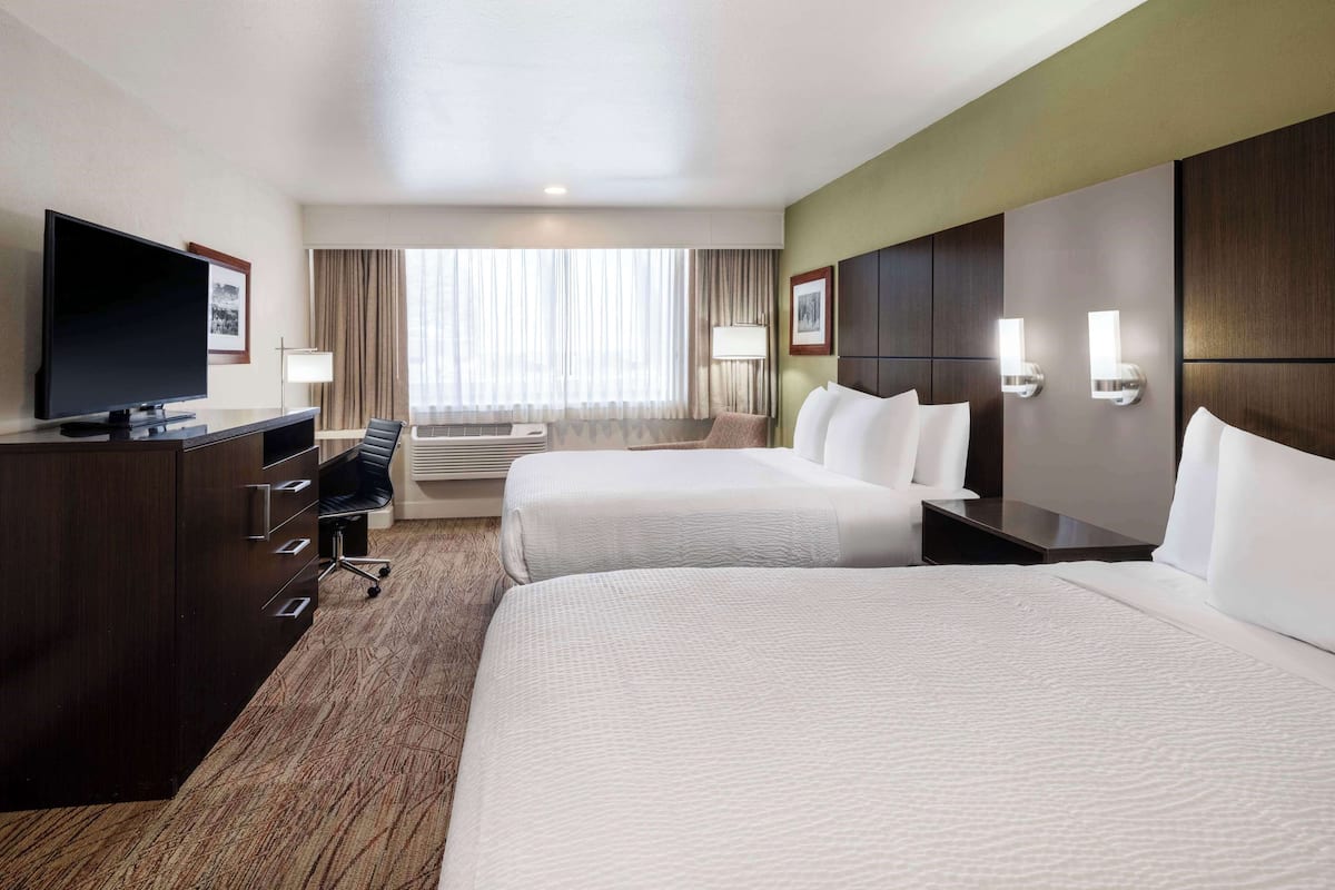 A hotel room with two beds, a desk with a flat-screen TV, a chair, a desk lamp, and a large window with curtains.