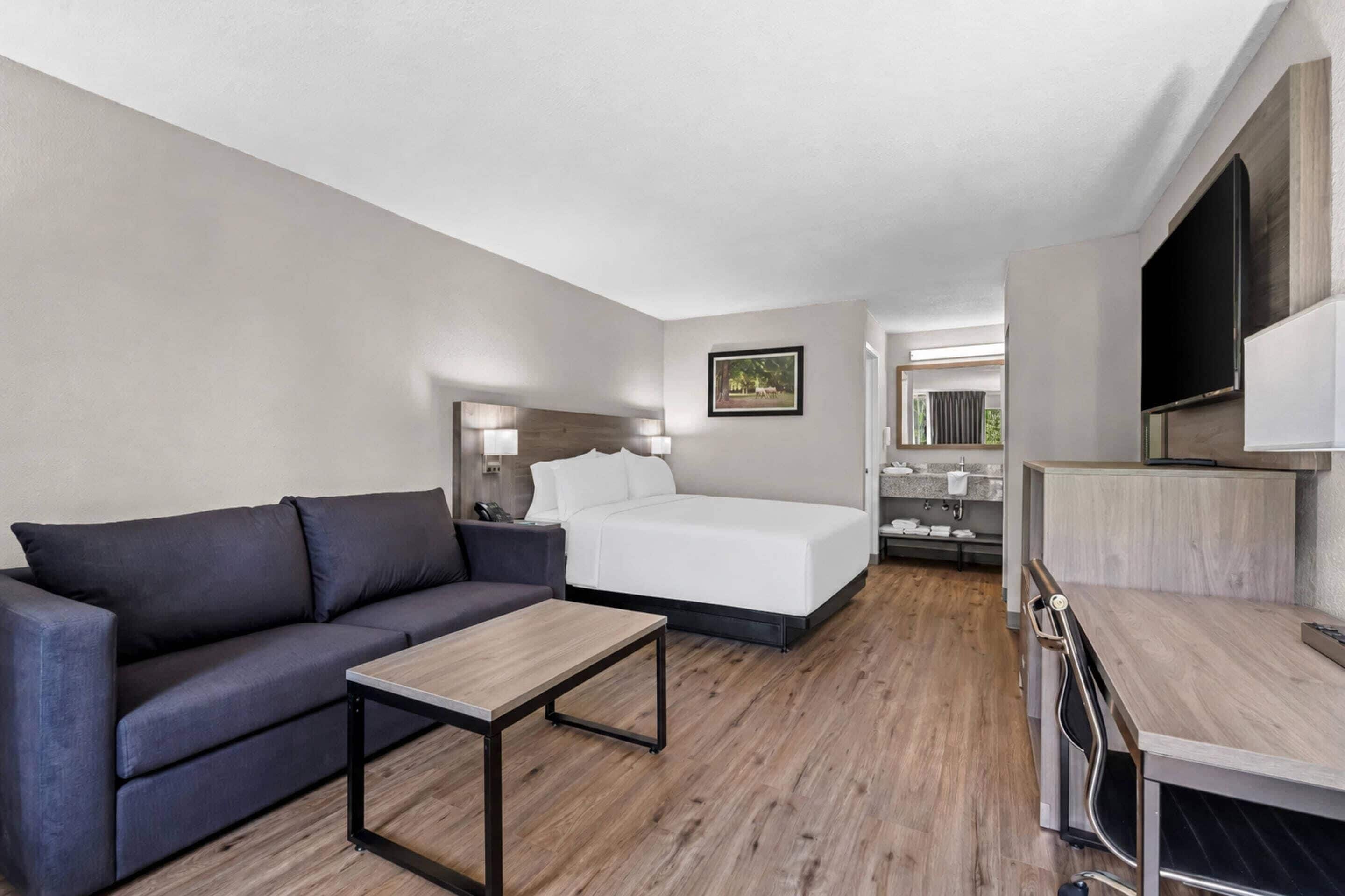 A hotel room with a bed, a sofa, a small table, a TV, and a bathroom visible through an open door.