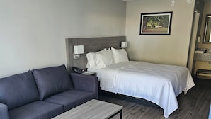 Habitación de hotel con un sofá gris, una mesa de centro de madera, una cama con ropa blanca y un cuadro en la pared.