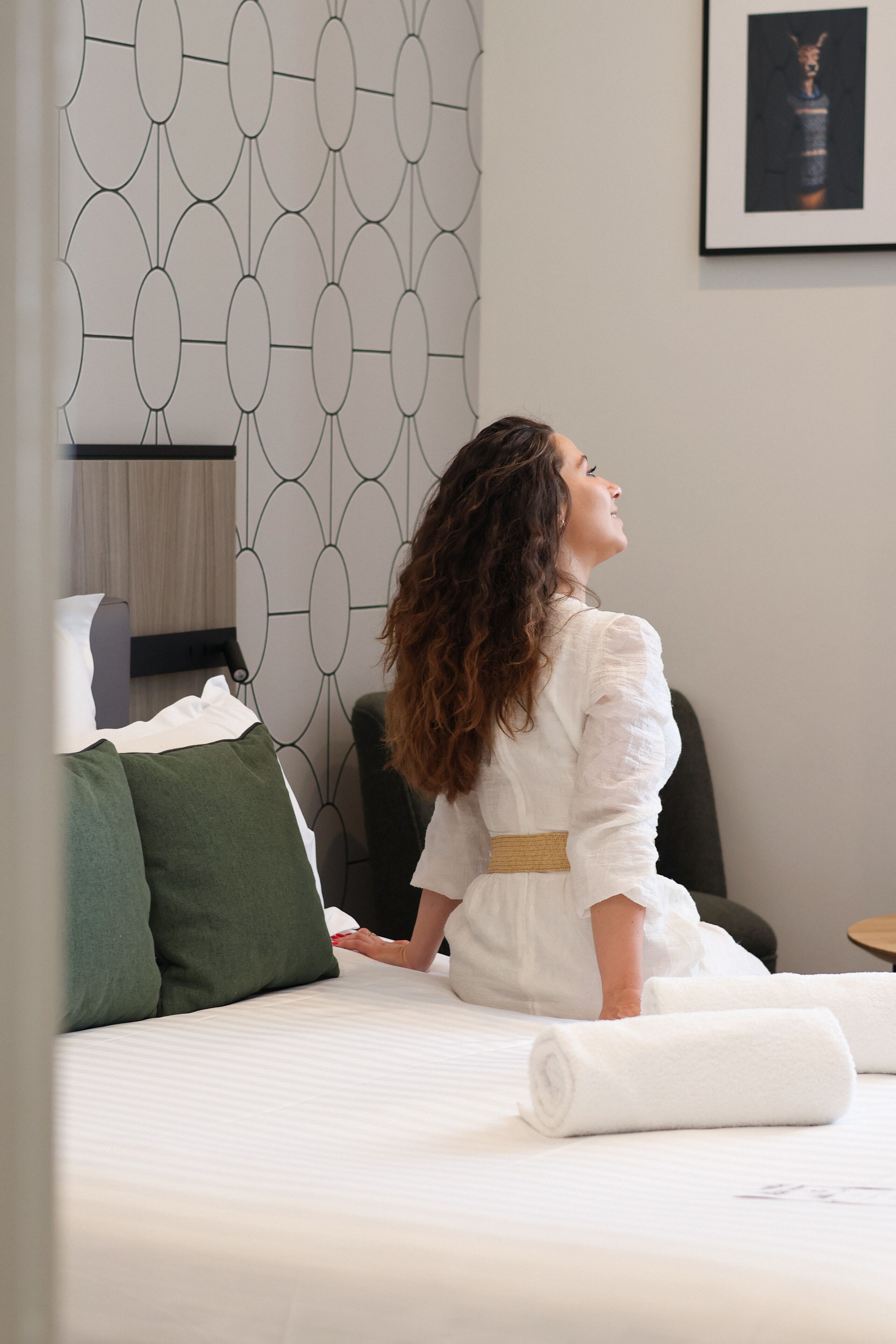 A woman sitting on a bed with a patterned wallpaper, a framed picture on the wall, and a rolled towel on the bed.