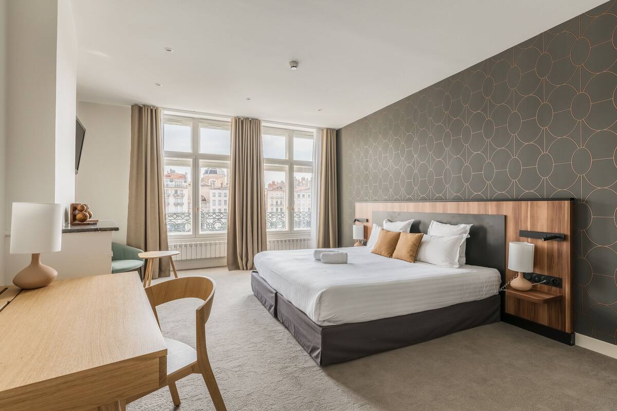 A modern hotel room with a large bed, a desk, a chair, and a view of the city through large windows.