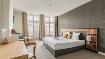 A modern hotel room with a large bed, a desk, a chair, and a view of the city through large windows.