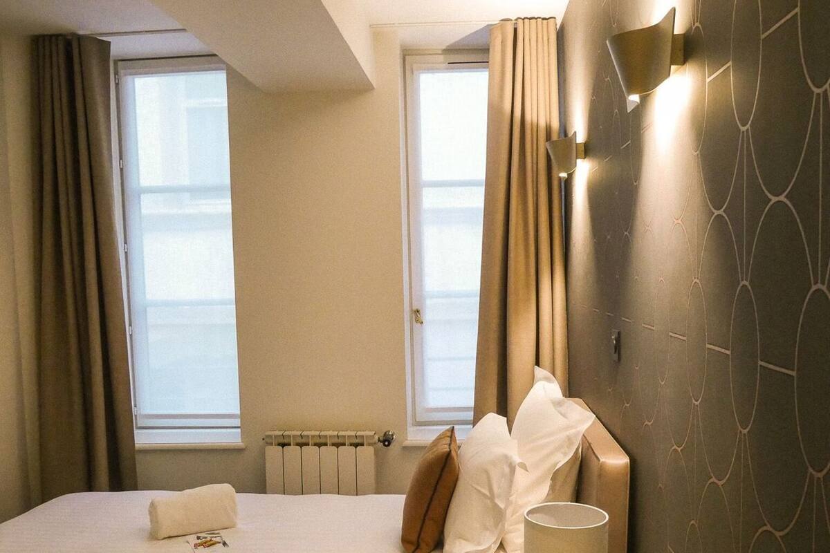 A hotel room with a bed, a bedside table with a lamp, a window with curtains, and a wall with a geometric pattern.
