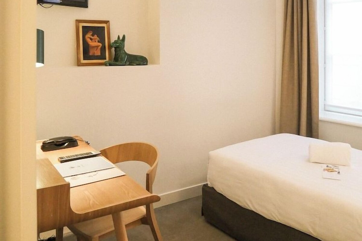 Habitación de hotel con cama, escritorio con silla, televisor montado en la pared y ventana con cortinas.
