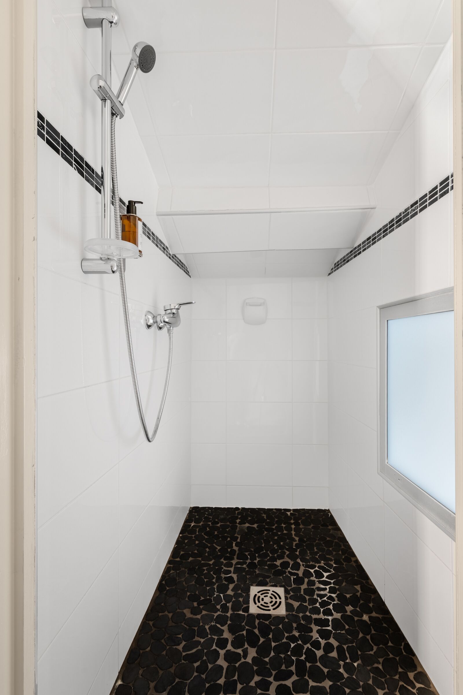 A small, modern shower room with white tiled walls and a black pebble-patterned floor.
