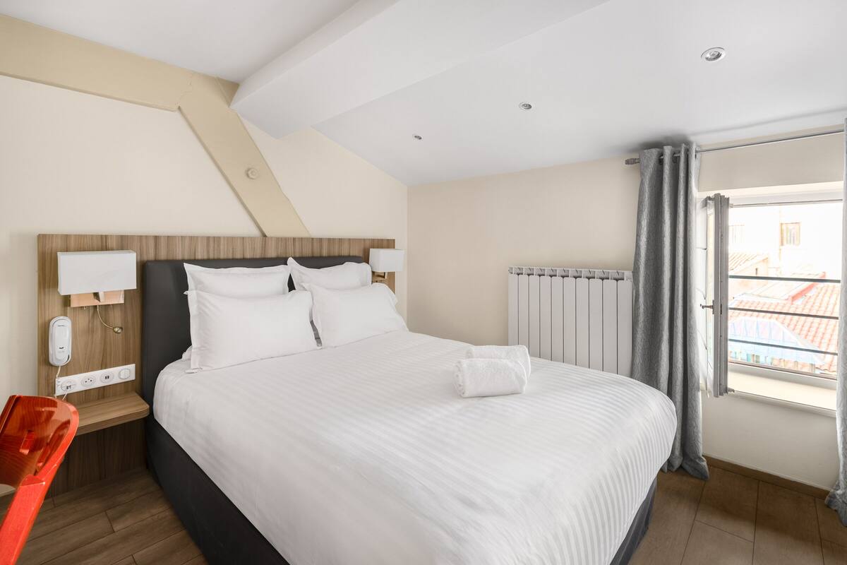 A hotel room with a large bed, bedside tables, a red chair, a window with curtains, and a view of buildings outside.