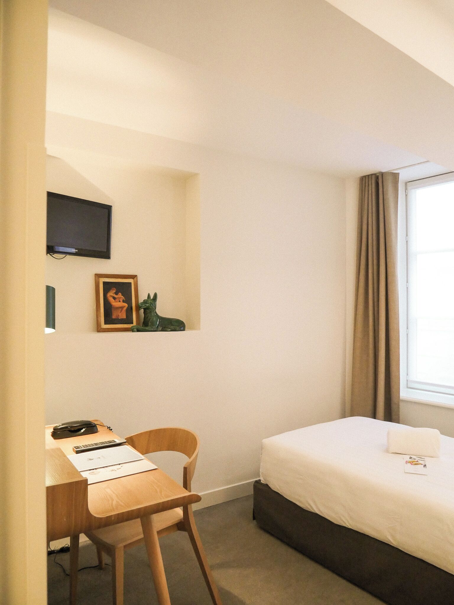 A hotel room with a bed, a desk with a chair, a television mounted on the wall, and a window with curtains.