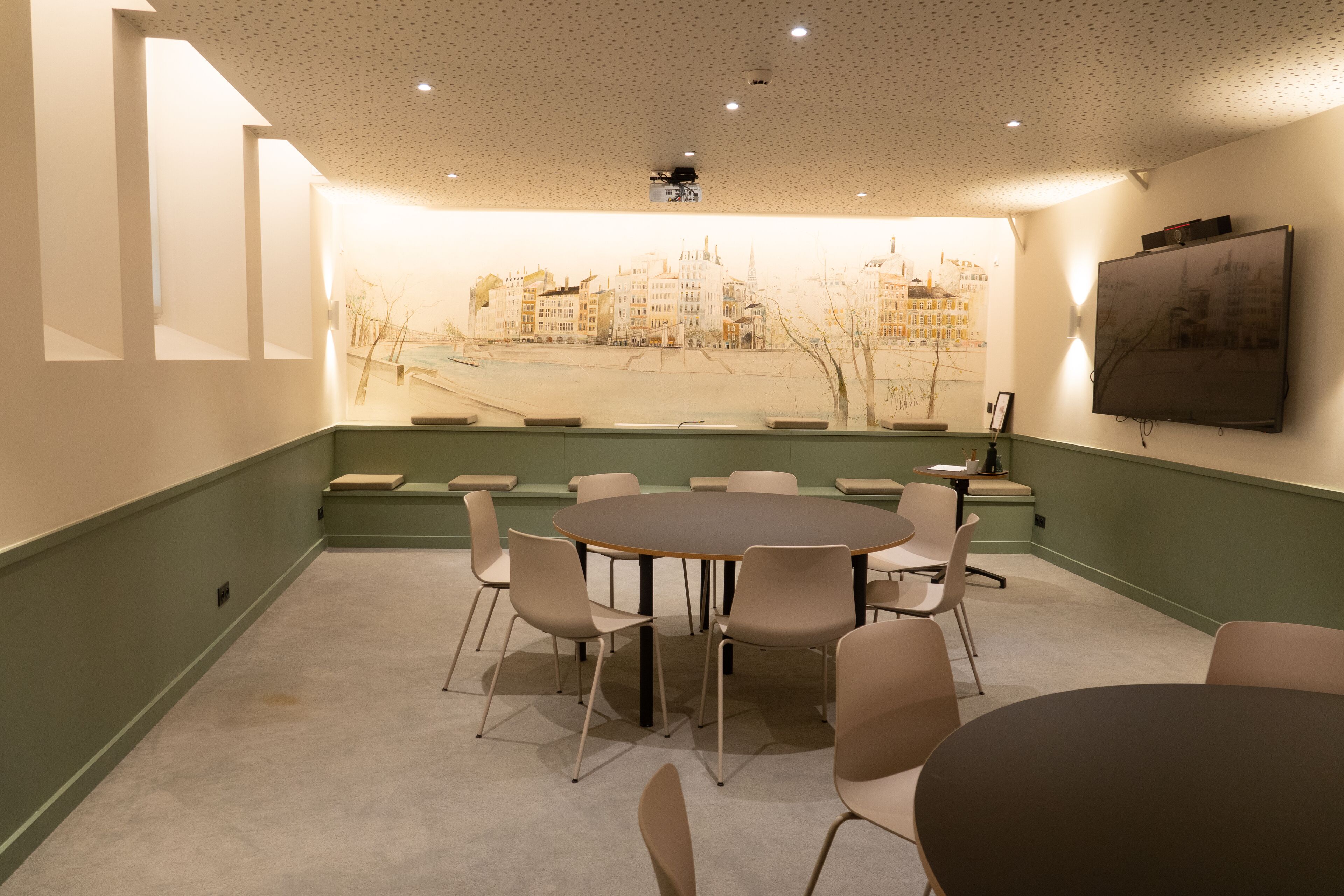A conference room with a large table surrounded by chairs, a projector screen, and a cityscape mural on the wall.