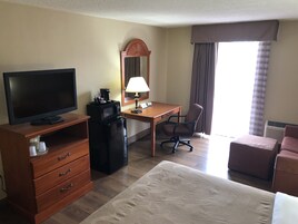 Habitación de hotel con televisión, escritorio con lámpara, una silla, cama y refrigerador pequeño.