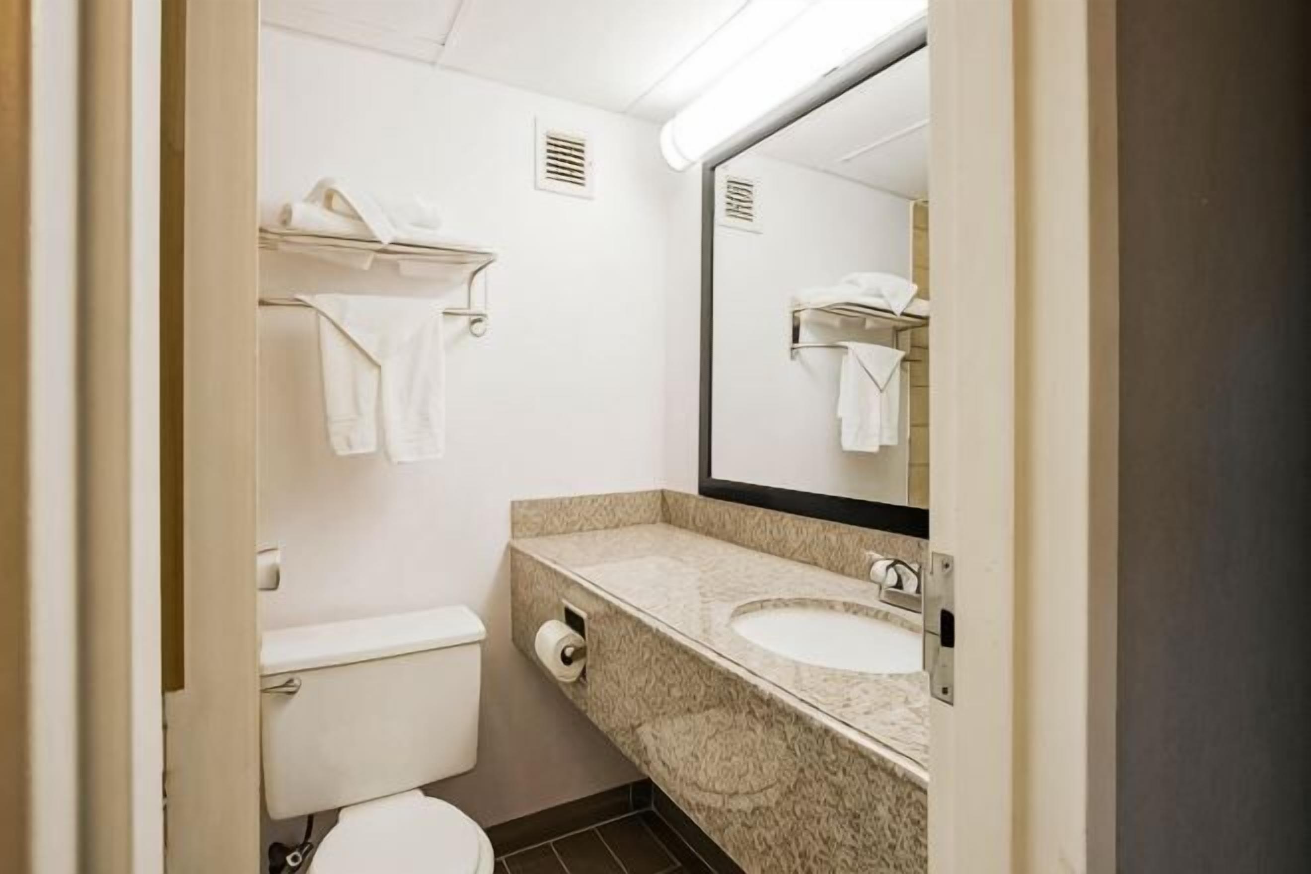 A bathroom with a toilet, sink, mirror, towel rack, and a towel dispenser.
