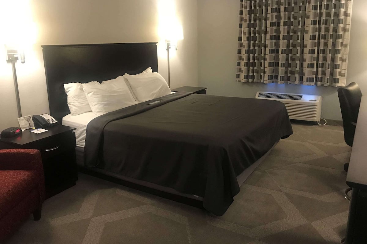 A hotel room with a bed, a nightstand with a phone and papers, a red chair, a desk with a lamp, and a window with patterned curtains.
