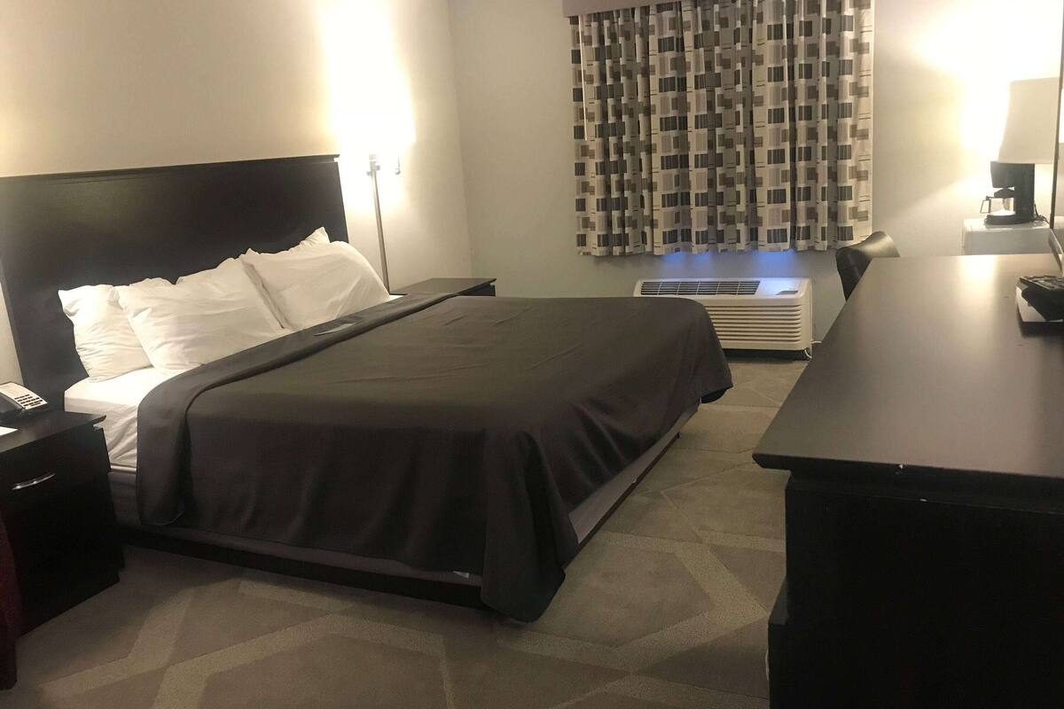 A hotel room with a bed, bedside table, lamp, air conditioning unit, and a desk with a chair.