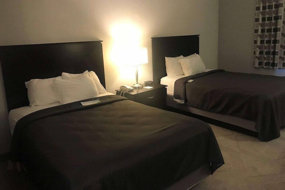 A hotel room with two beds, each with a dark headboard and white bedding. There is a nightstand with a lamp and a phone between the beds.