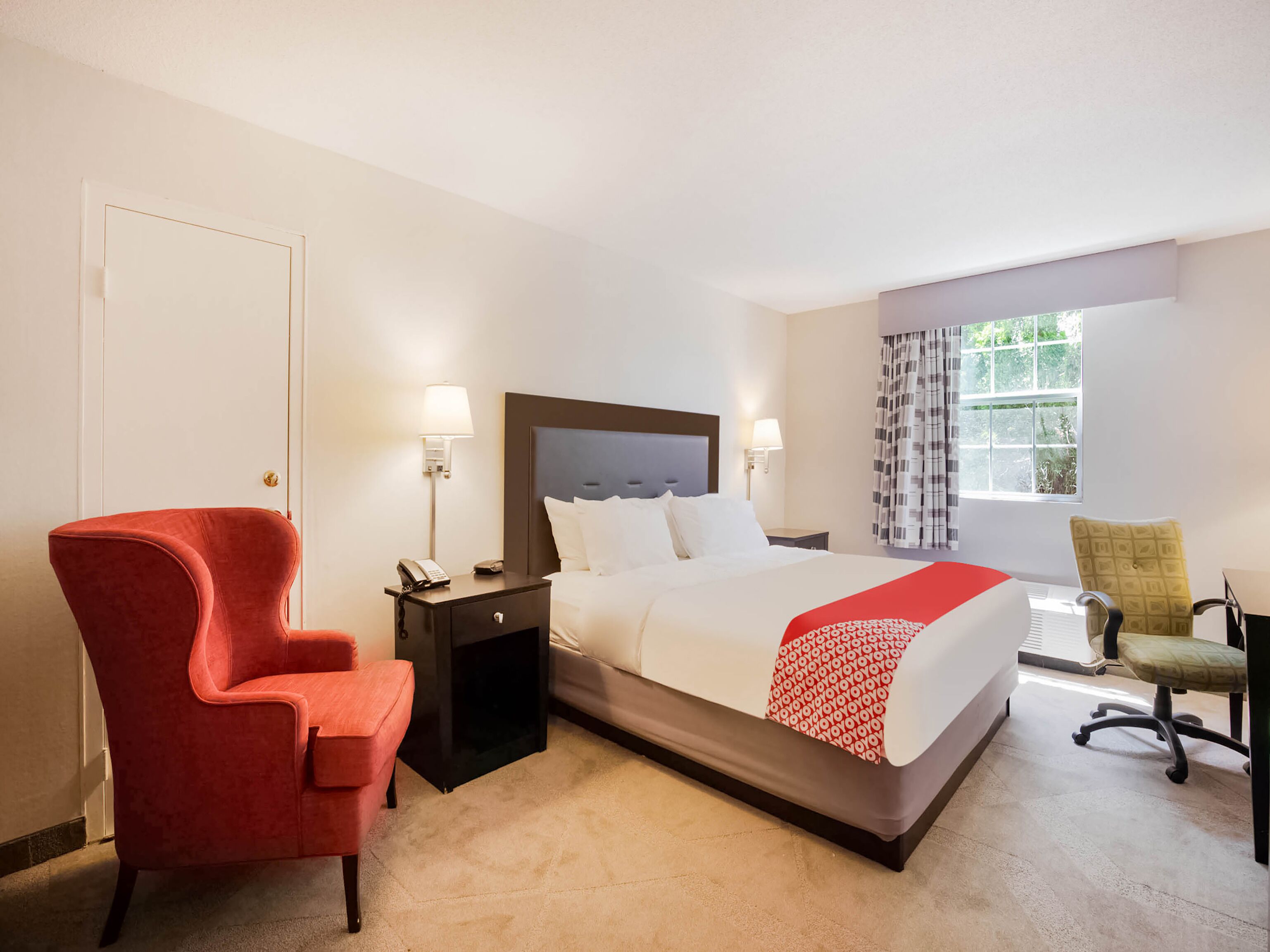 A hotel room with a bed, bedside tables, a red armchair, a green office chair, and a window with curtains.