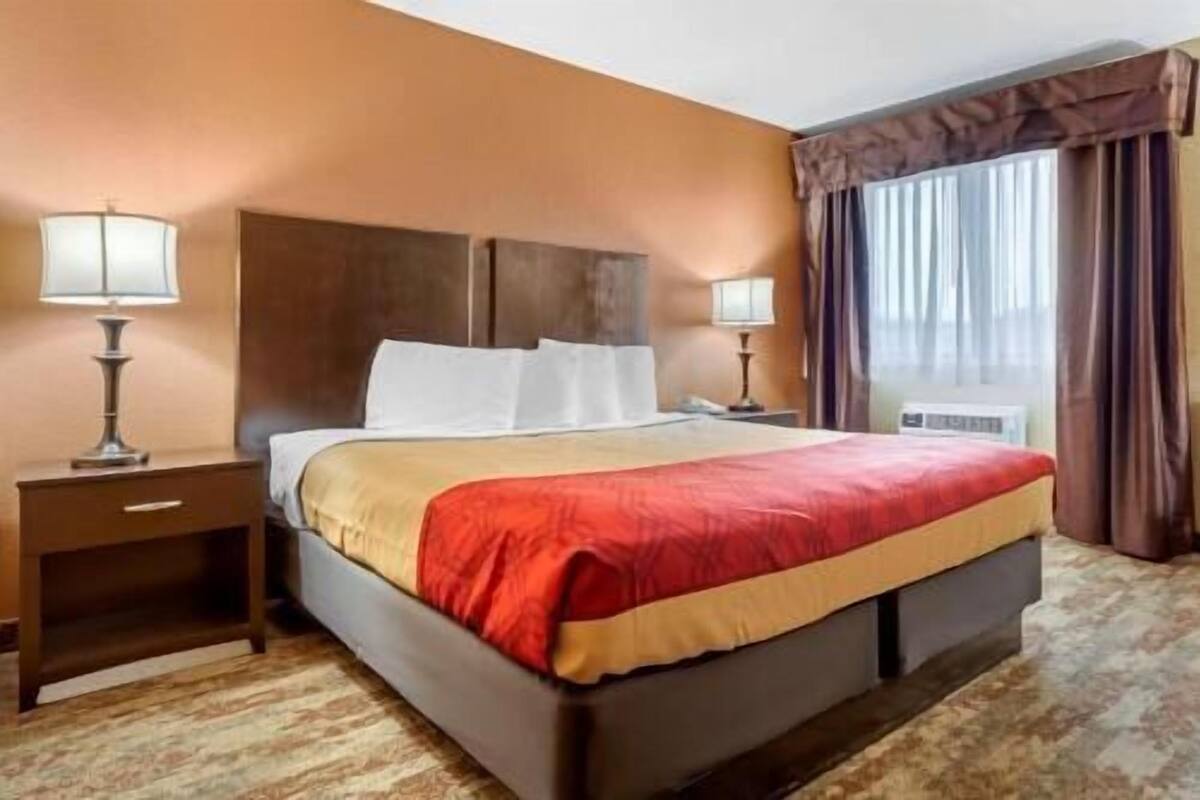 Une chambre d’hôtel avec un lit, deux tables de chevet, une lampe sur chaque table, une fenêtre avec des rideaux et un sol recouvert de moquette.