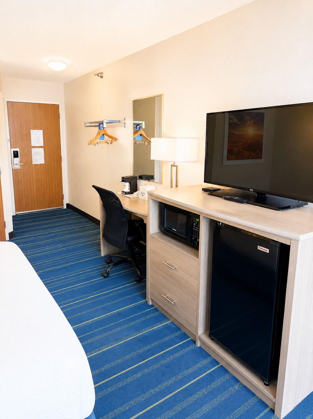 Quarto de hotel com uma cama, uma escrivaninha com computador, micro-ondas, televisão e um pequeno frigorífico.
