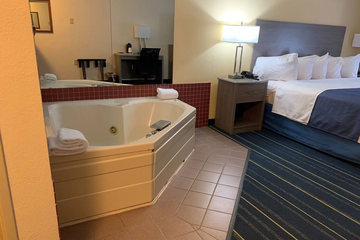 A hotel room with a large bathtub, a bed with white linens, a bedside table with a lamp, and a small desk with a chair.