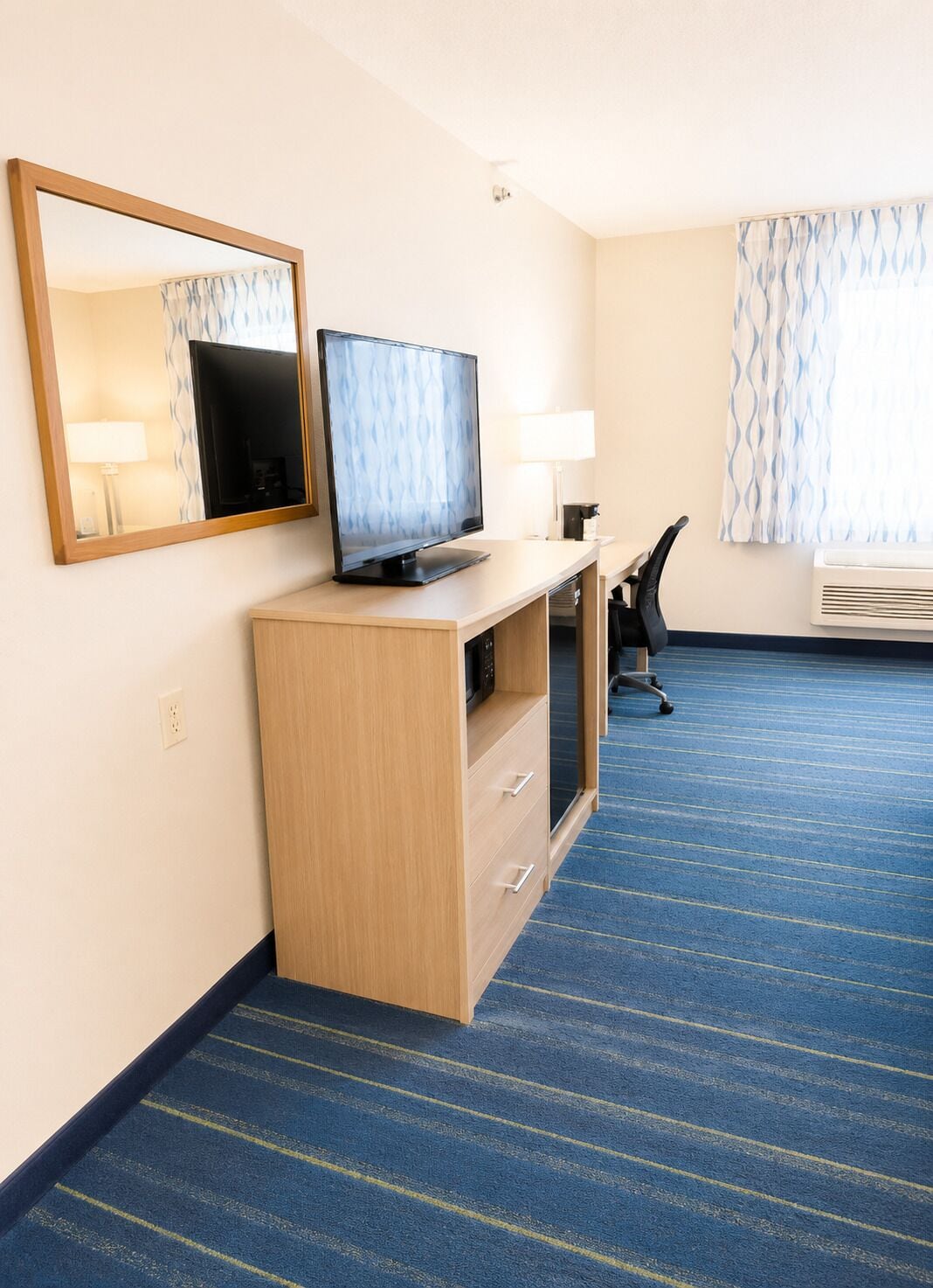 A hotel room with a desk, a television, a mirror, a chair, and a window with curtains.