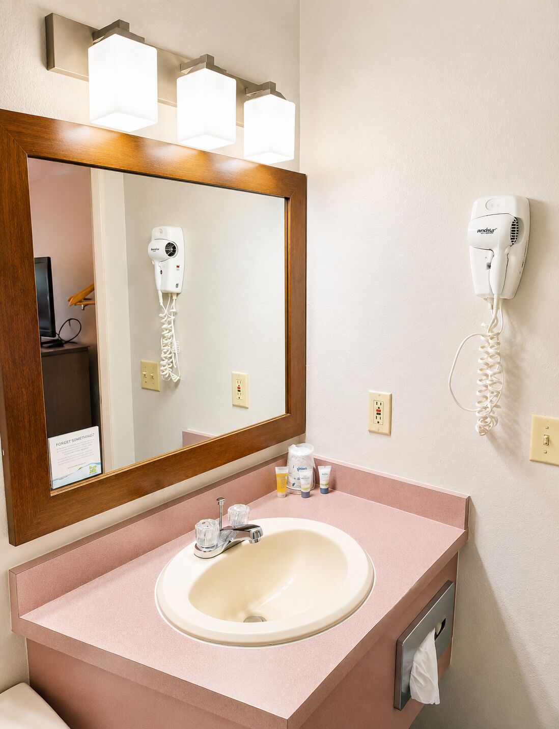 A bathroom with a sink, mirror, hairdryer, and a towel dispenser.